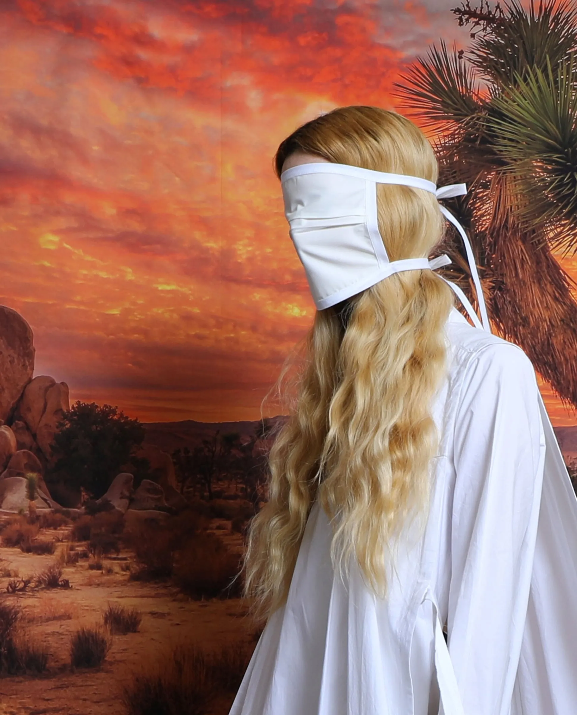 Woman with long blonde hair in a white robe and white face covering, seen from behind against a red desert sky