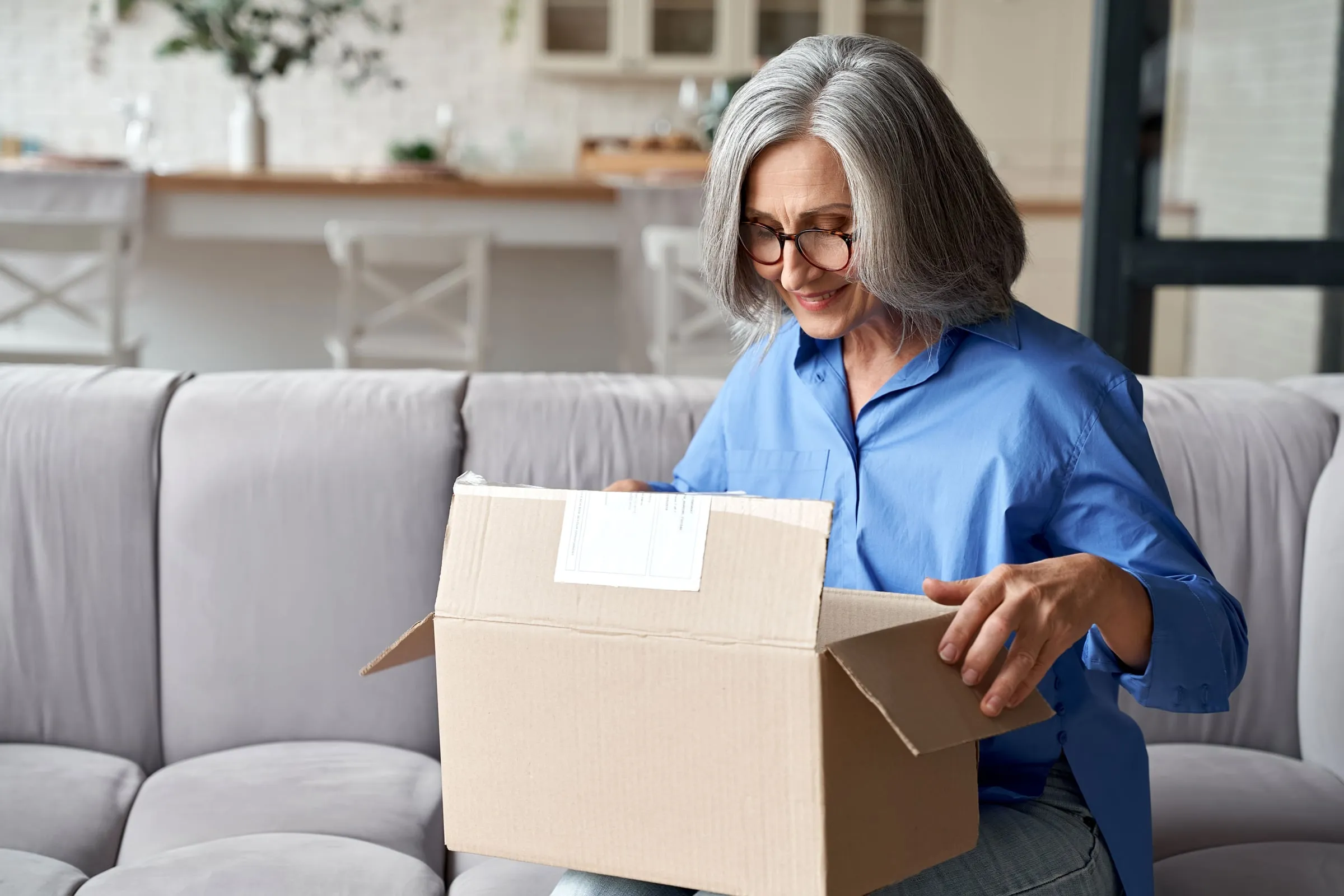 Woman with gray hair in a blue shirt opening a cardboard box on a sofa