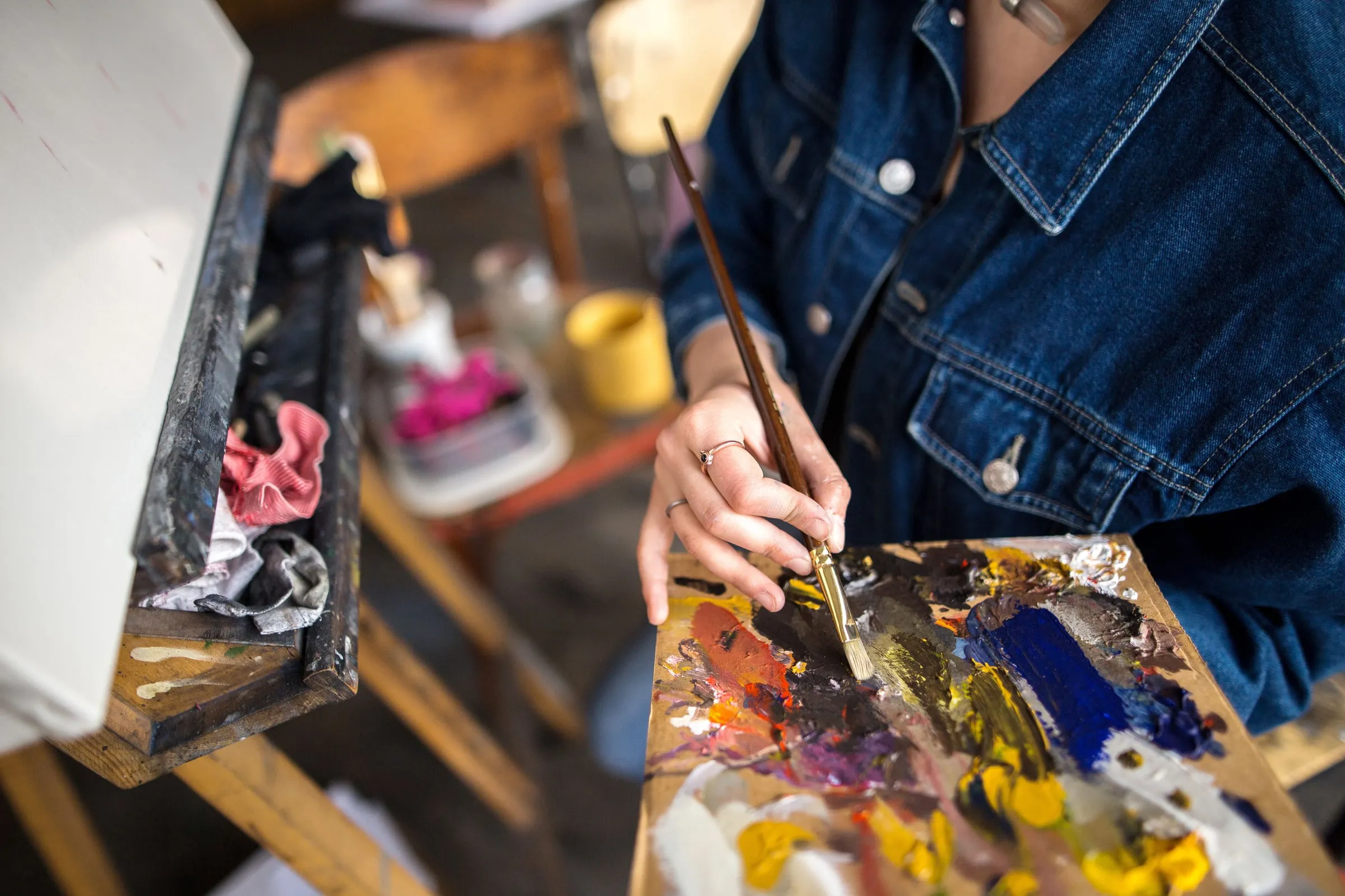 Artist in a denim jacket painting at an easel, holding a brush over a paint-covered palette